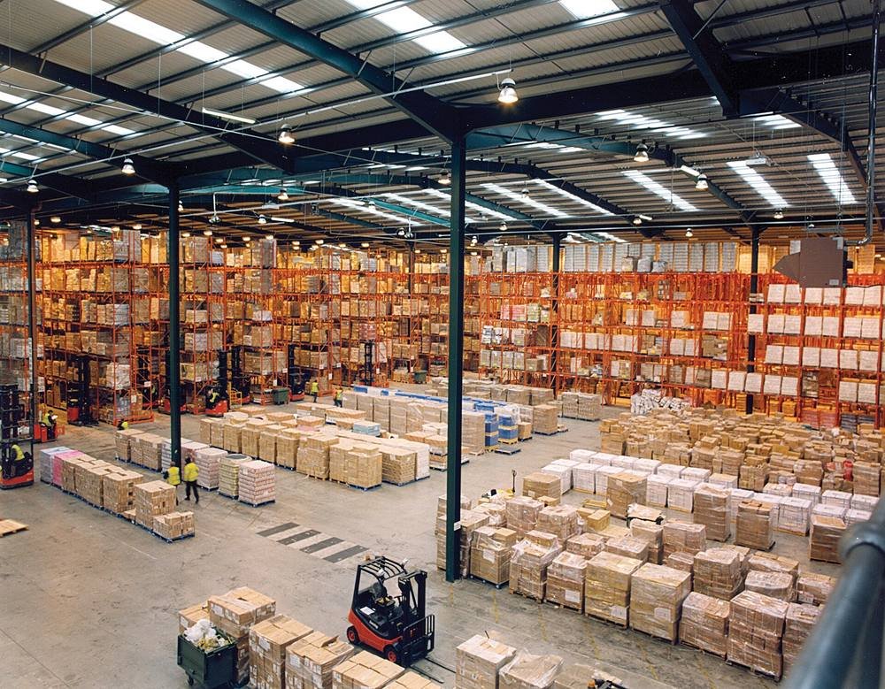 Interno di un magazzino logistico: scaffalature porta-pallet su più livelli, merce pallettizzata e carrelli elevatori in attività.